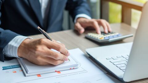 Im Bild ist der Oberkörper eines Mannes zu sehen, der auf einem Block Notizen anfertigt. Auf dem Tisch sind zudem ein Laptop und Arbeitsunterlagen sichtbar. 