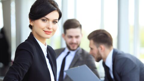 Im Vordergrund lehnt eine junge, lächelnde Frau mit Sakko und Bluse an einem Tisch in einem Büro. Im Hintergrund unterhalten sich zwei junge Kollegen.