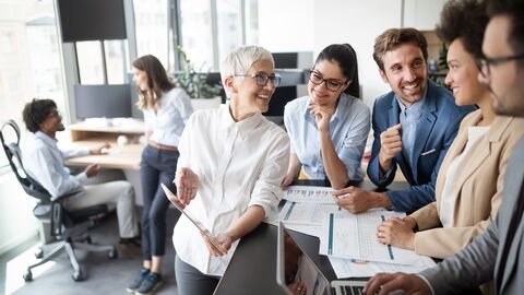 Im Vordergrund steht ein Team an einem Schreibtisch und unterhält sich über einen Sachverhalt. Die Stimmung ist gut. Im Hintergund ist ein Büro mit weiteren Mitarbeitenden zu sehen.