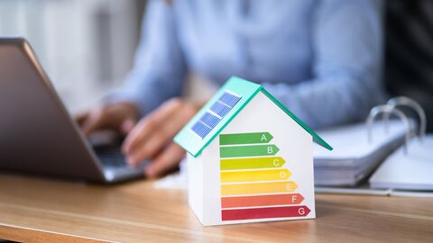 Beim LBIH werden die Aufgaben des Energiemanagements in einem Competence-Center Energie (CC Energie) gebündelt. Im Vordergrund steht ein kleines Haus mit Solardach auf einem Schreibtisch. Auf dem Haus sind die Energieeffizienzklassen dargestellt. Im Hintergrund ist unscharf eine arbeitende Person an einem Laptop zu erkennen.