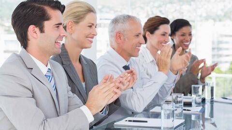 In einem Büro sitzen fünf lächelnde Teammitglieder an einem langen Glastisch und applaudieren.