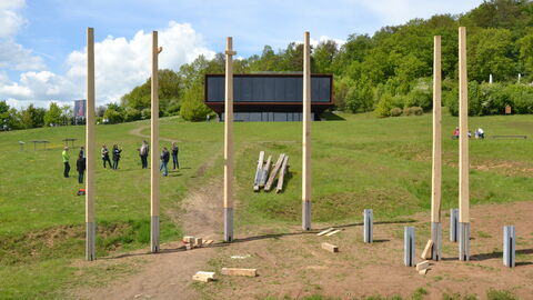 Keltenweld am Glauberf Sechs Eichenpfosten auf einer Wiese mit Personen und einem Gebäude im Hintergrund