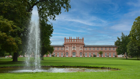 Vor dem Schloss Biebrich in Wiesbaden erstreckt sich der Schlosspark mit einer großen Rasenfläche, auf der ein Brunnen mit Wasserfontäne zu sehen ist. Das Bild ist eingerahmt vom alten Baumbestand im Schlosspark Biebrich.
