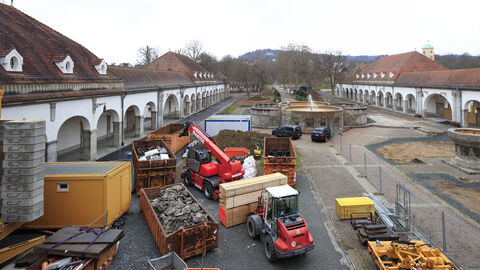 Baustelle mit verschiedenen Fahrzeugen, Containern und Materialien in einem Innenhof mit Springbrunnen 