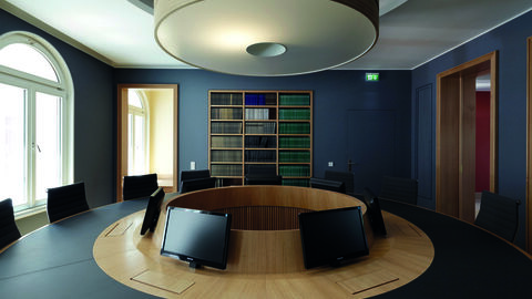 Die besondere Gestaltung des Staatsgerichtshofs verbindet Würde und Bescheidenheit des Staatsorgans mit dem Stolz, Hüter der Hessischen Verfassung zu sein Im Bild ist das Beratungszimmer der Richter im Hessischen Staatsgerichtshof in Wiesbaden zu sehen. Im Vordergrund steht ein runder Tisch mit Monitoren. Die satte dunkelblaue Farbe der Wand sowie verschiedene Holzelemente an Tisch, Regal und Türrahmen geben dem Raum seinen Charakter.